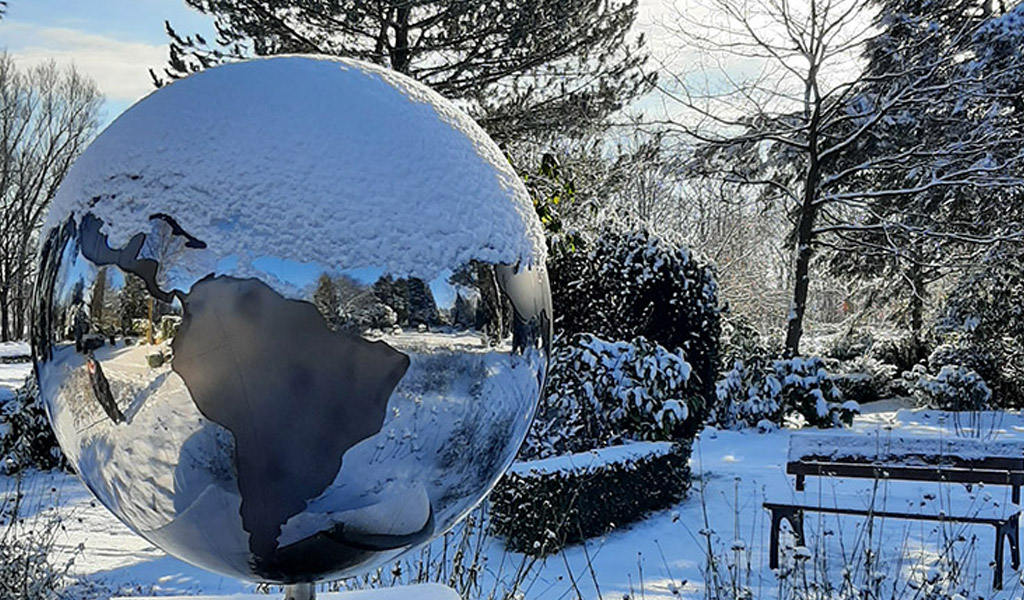 Friedhof Blumenbeet Weltkugel Urne Spiegel im Winter mit Bank und Bäume im Schnee