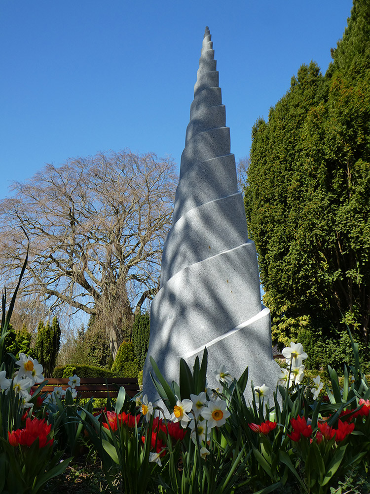 Friedhof: Blumenbeet mit Kunstwerk Himmelsleiter vor Bäumen und Himmel hinter bunten Blumen